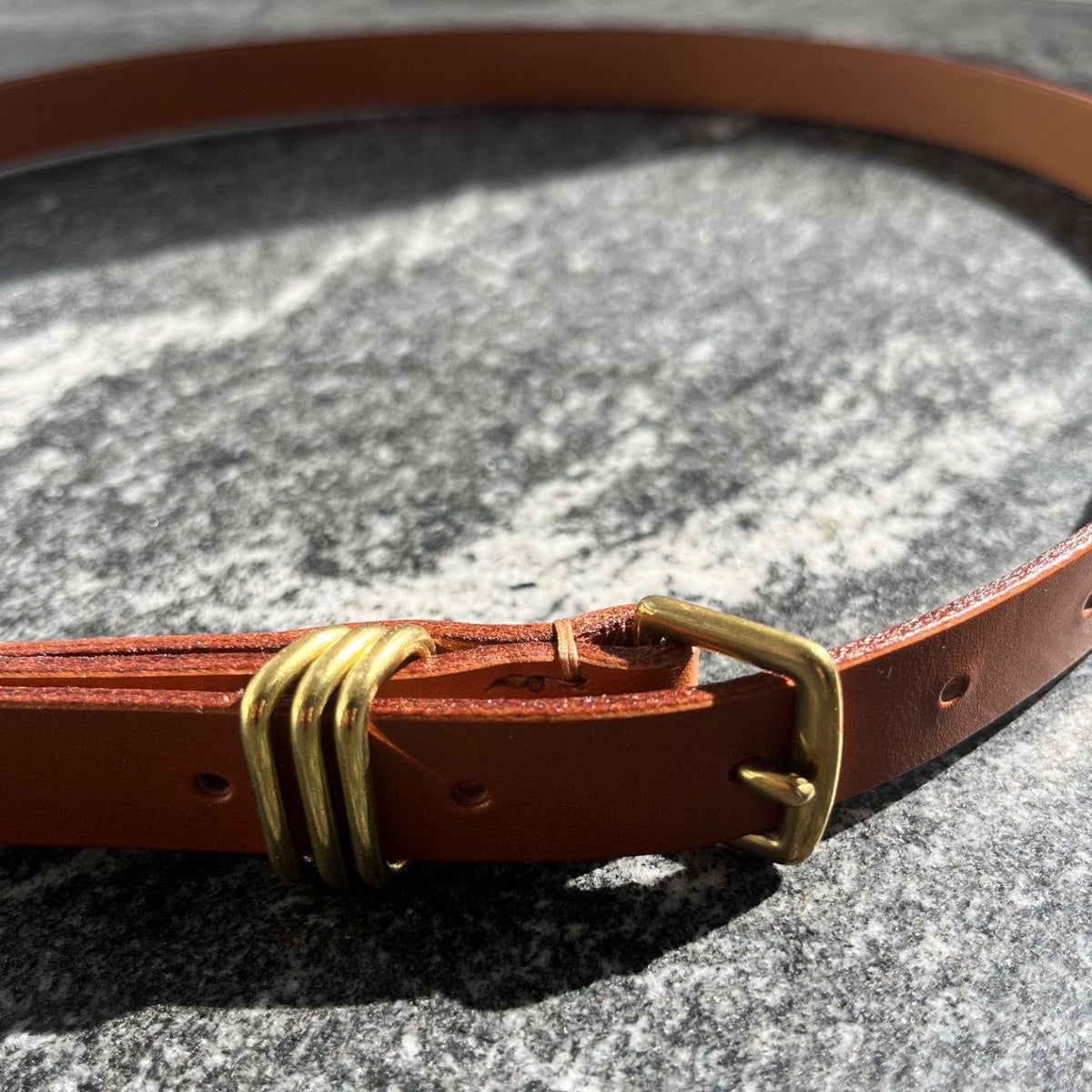 A close-up of a tan leather belt with a brass buckle and a decorative metal loop detail, resting on a textured gray stone surface.