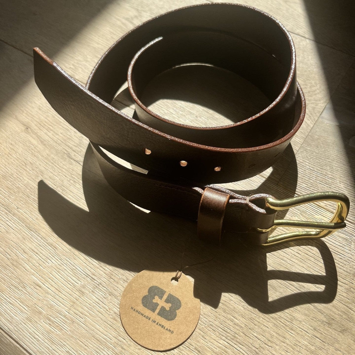 A dark brown leather belt with a brass buckle, coiled on a light wood floor with dramatic sunlight and shadows. A round tan paper tag with a dark logo lies in front of the belt.
