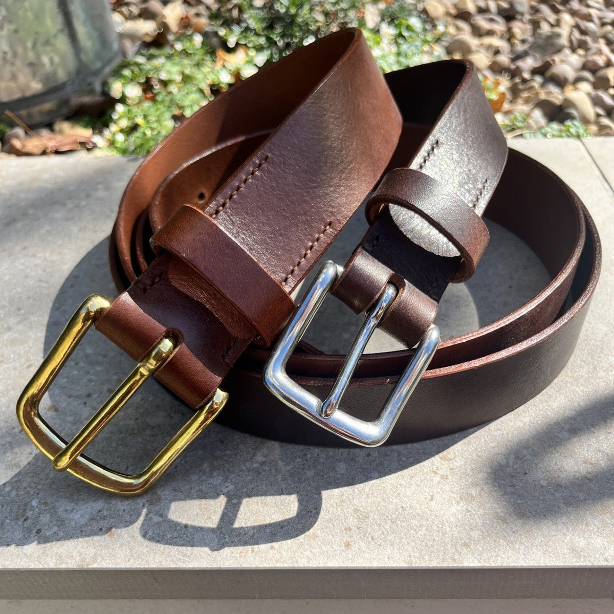 Two leather belts, one brown with a gold buckle and one dark brown with a silver buckle, coiled on a stone surface outdoors.