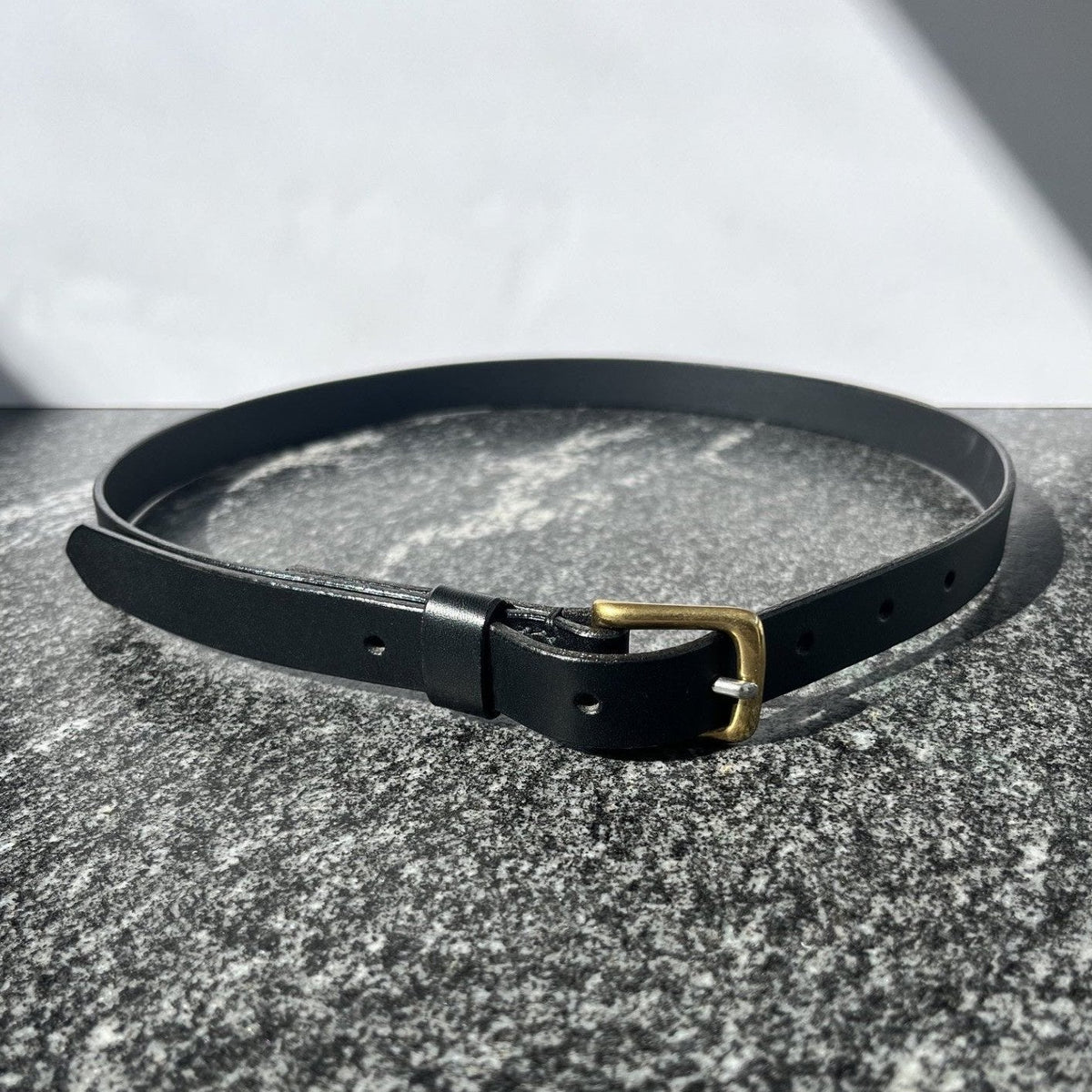 A black leather belt with a gold-tone buckle, coiled on a textured gray stone surface.