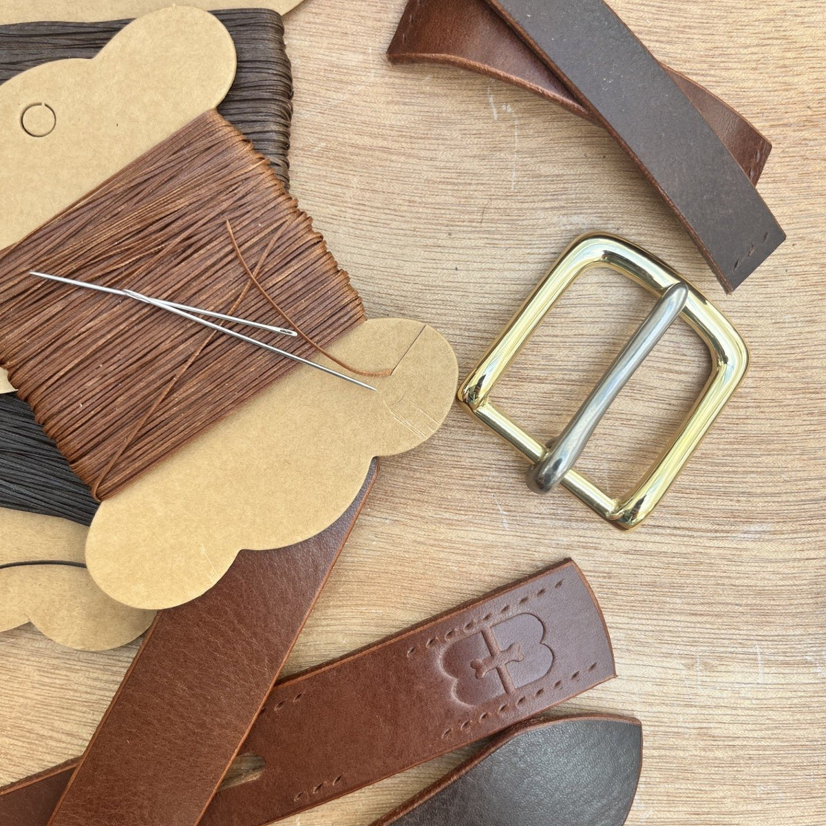 A flat-lay of leather crafting materials on a light wood surface, including brown and dark brown leather strips, a spool of brown thread, a brass buckle, and a needle.