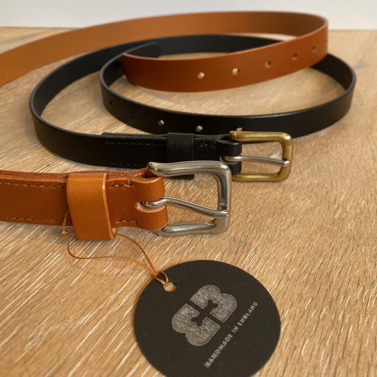 Two leather belts in tan and black arranged on a light wood surface with a round black brand tag hanging from the tan belt. The belts have brass and silver-tone buckles.
