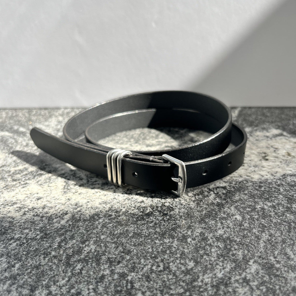 A black leather belt with a silver-toned metal buckle, coiled on a textured gray surface against a light gray background.