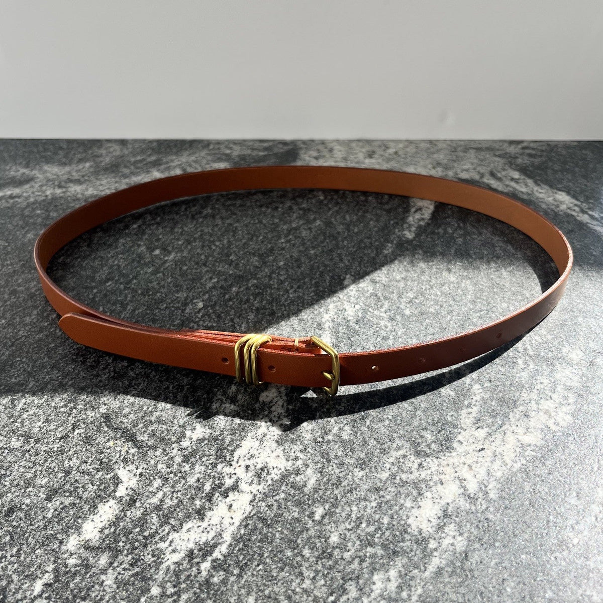 A slim tan leather belt with a gold coloured buckle, displayed in a circular shape on a textured gray stone surface against a light gray background.