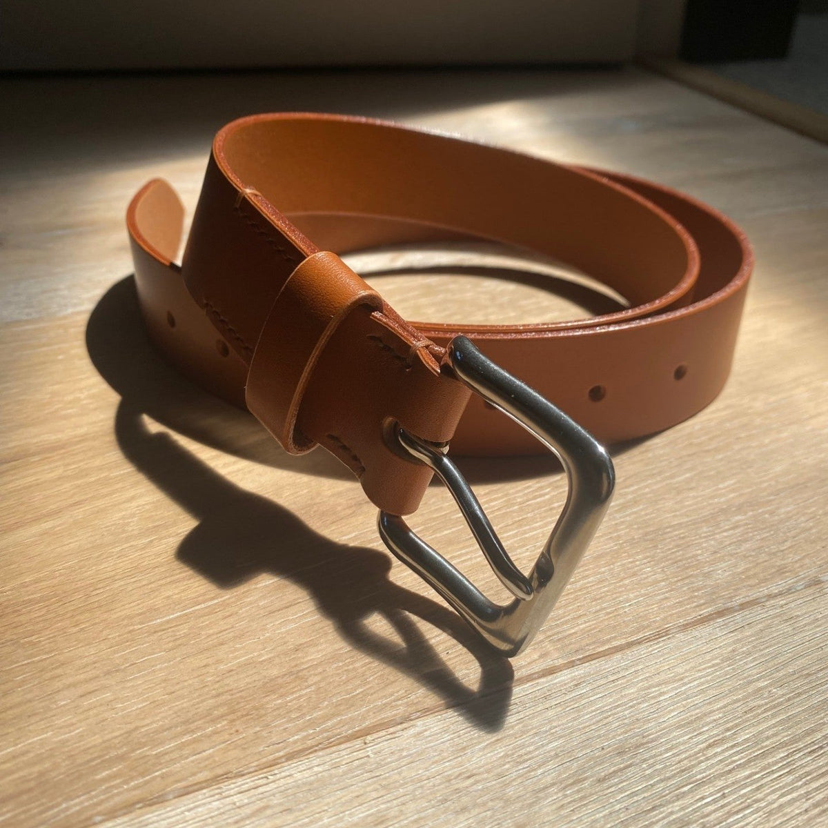 A tan leather belt with a silver metal buckle, coiled on a light wood surface under warm, direct sunlight that casts a sharp shadow.