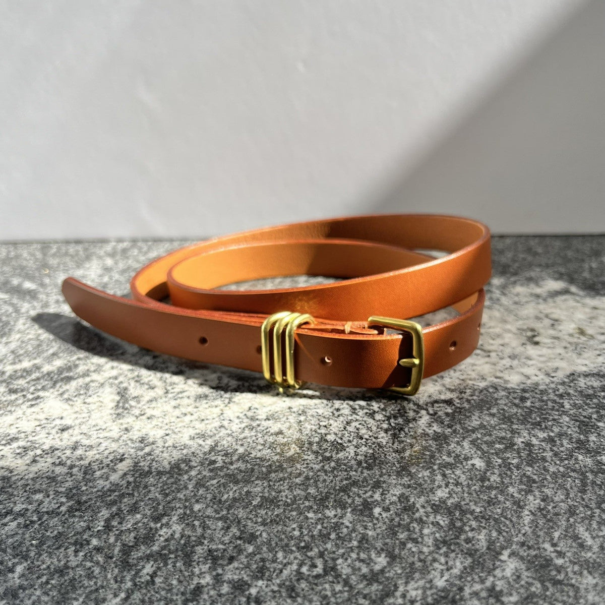 A warm tan leather belt with a gold-toned triple closure, coiled on a textured gray stone surface against a neutral light gray background.
