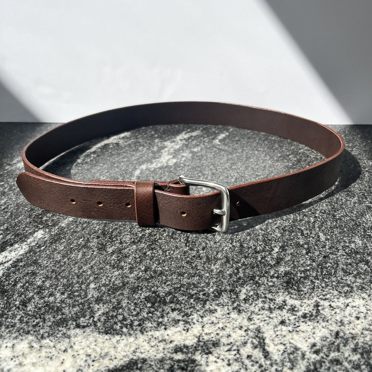 A brown leather belt with a silver metal buckle, arranged in a loop on a textured gray stone surface with a white background and diagonal shadows.