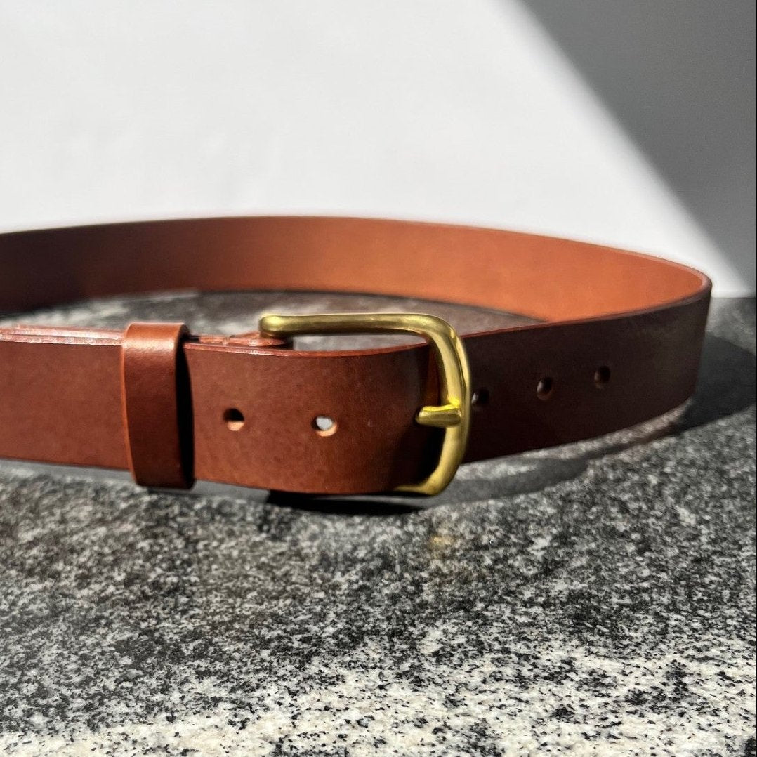 A brown leather belt with a brass metal buckle, displayed on a textured gray stone surface with a white background.