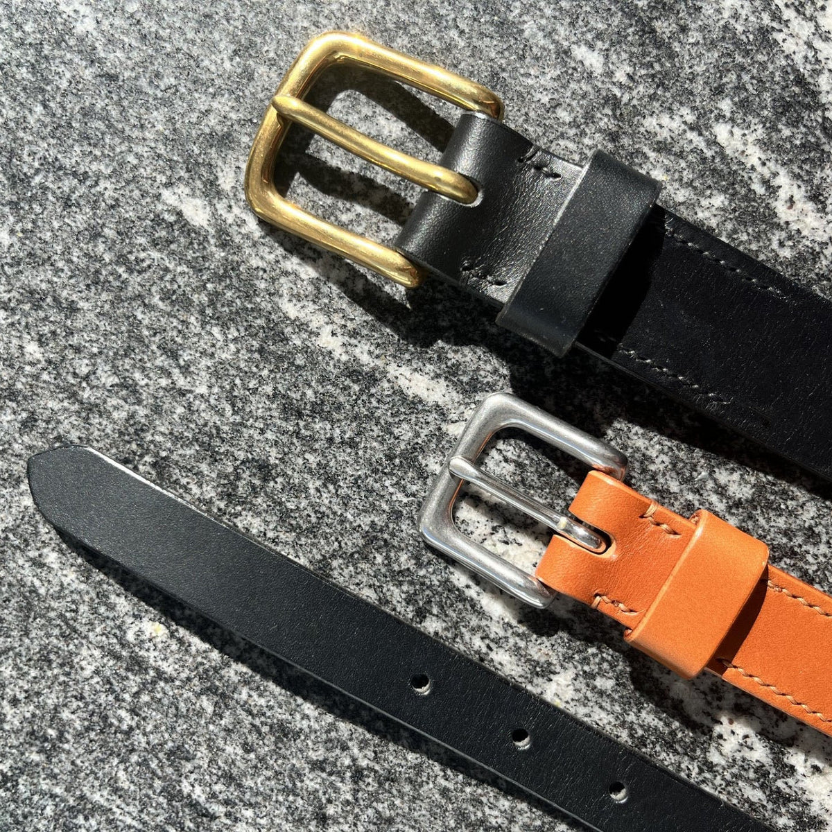 Two leather belts laid diagonally on a textured gray stone surface. One belt is black with a brass buckle, and the other is tan with a silver-tone buckle.