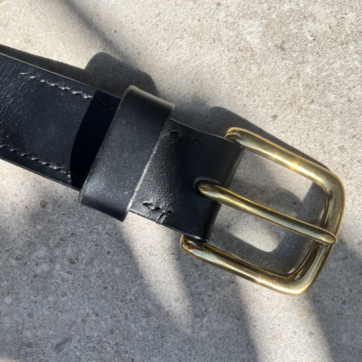 A close-up of a black leather belt with a polished brass buckle, laid diagonally on a textured gray concrete surface with natural shadows.
