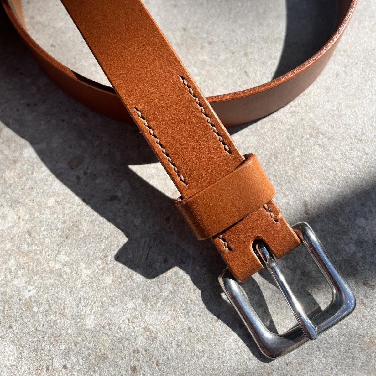 A close-up of a tan leather belt with a silver metal buckle, against a textured concrete background.