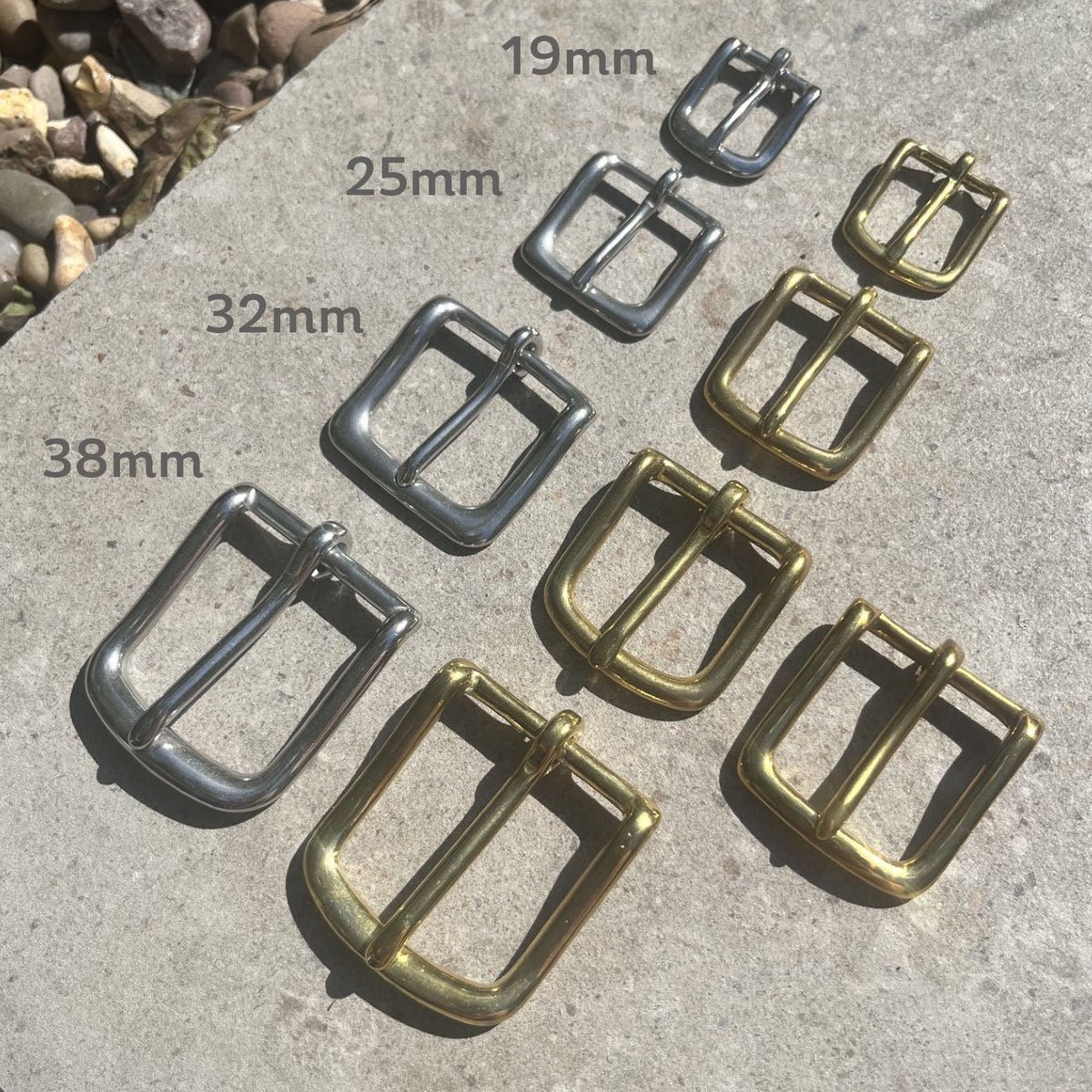 Nine brass and silver metal belt buckles arranged on a concrete surface with size labels 19mm, 25mm, 32mm, and 38mm overlaid next to each buckle.