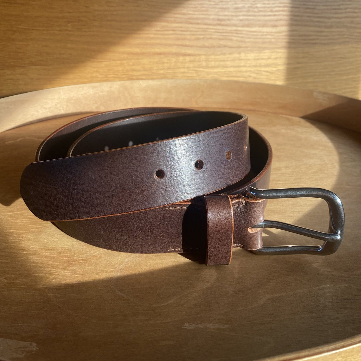 A dark brown textured leather belt with a silver metal buckle, coiled on a wooden tray with warm sunlight casting shadows.