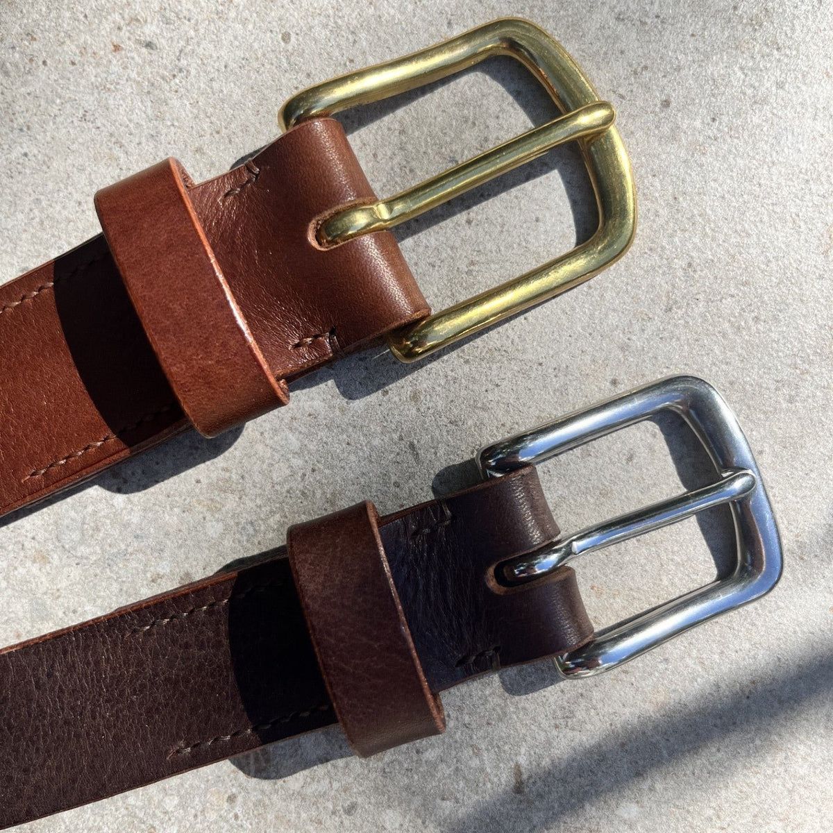 Two leather belts with brass and silver buckles laid diagonally on a light concrete surface.