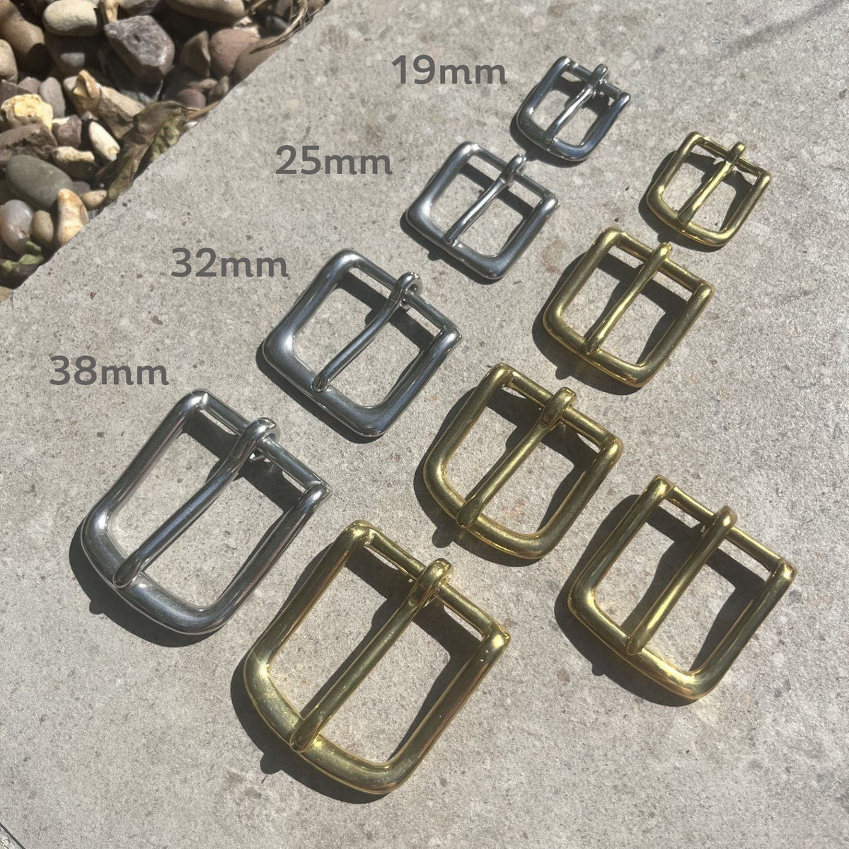 Nine brass and silver metal belt buckles arranged on a concrete surface with size labels 19mm, 25mm, 32mm, and 38mm overlaid next to each buckle.