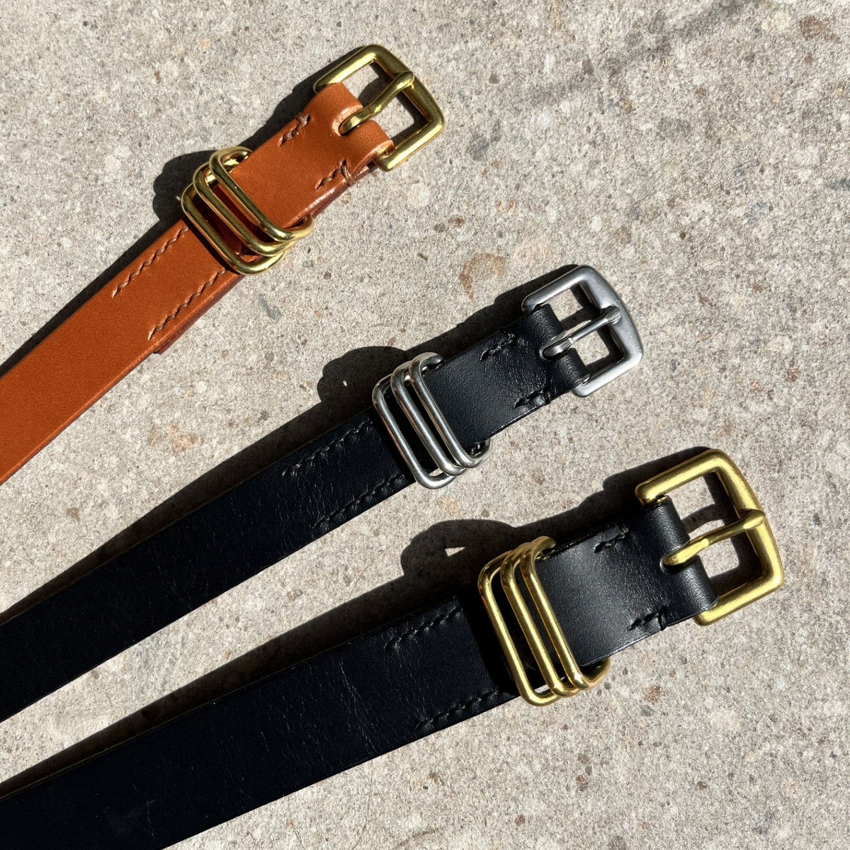 Three belt straps laid diagonally on a textured concrete surface. The top belt is tan with a brass buckle, and the two lower belts are black with silver and brass buckles respectively.