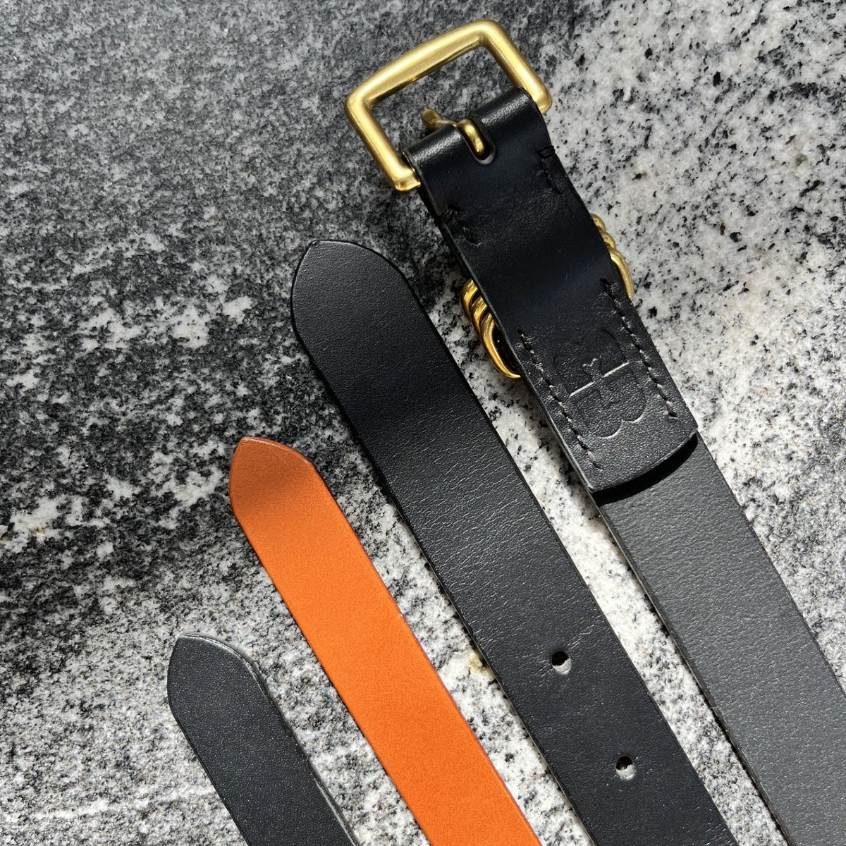 A close-up flat lay of leather belts on a textured gray surface, showing a black sbelt with a gold buckle, a black belt, and a tan belt