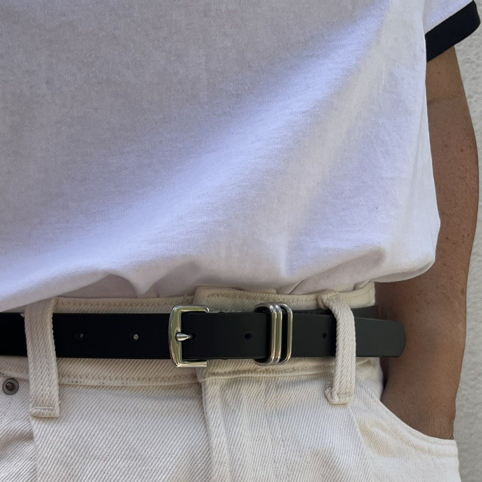 A close-up of a black leather belt with a silver-toned metal buckle worn over cream-colored trousers and a white t-shirt.