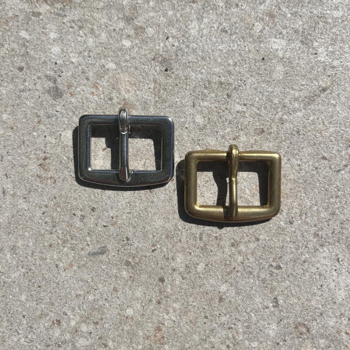 Two metal belt buckles, one silver-toned and one gold-toned, placed on a textured gray concrete surface.
