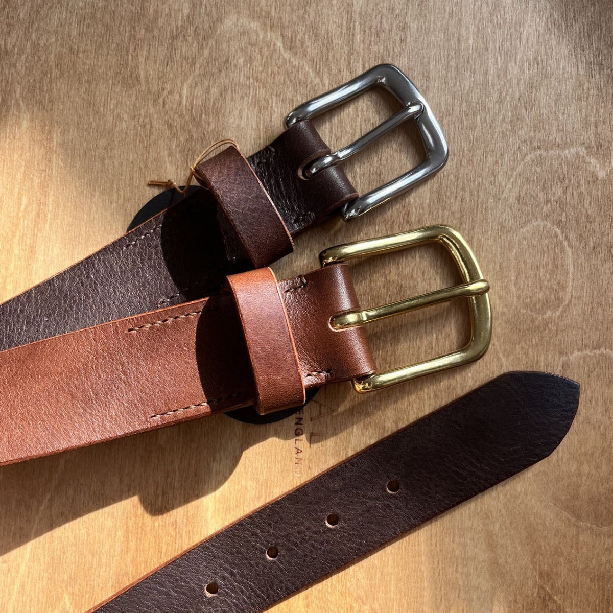 Two leather belts in tan and dark brown tones with silver and brass buckles, arranged on a textured wooden surface.