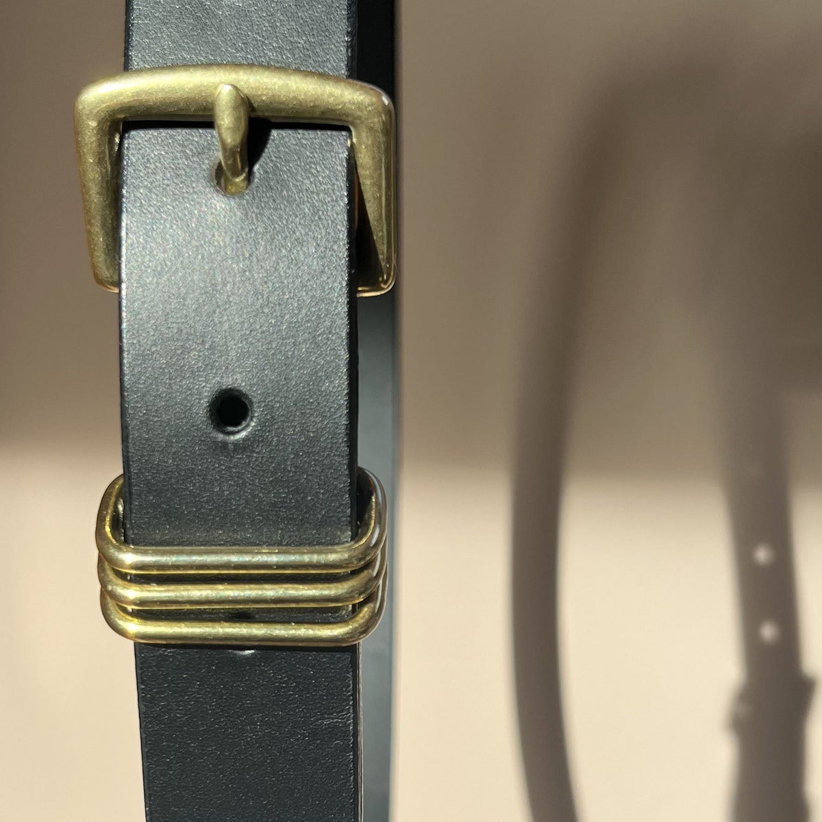 A close-up of a black leather belt with  brass hardware, including a buckle and a triple-ring keeper, set against a warm beige background with a soft shadow.