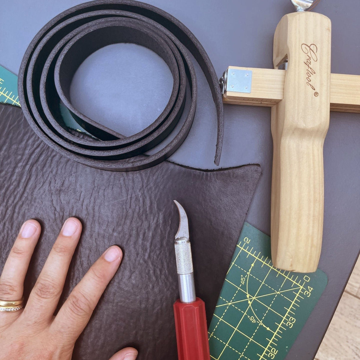 A leatherworking setup on a gray table showing a rolled dark black leather strip, a large piece of black leather, a wooden strap cutter with a visible Craftsman logo, a red-handled cutting tool, and a green self-coping cutting mat. A person's hand is visible holding the leather piece.
