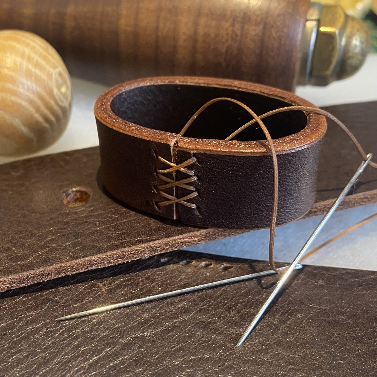A close-up of a hand sewn dark brown leather belt loop shown with leatherworking tools including a wooden mallet, leather strip, and metal needles.