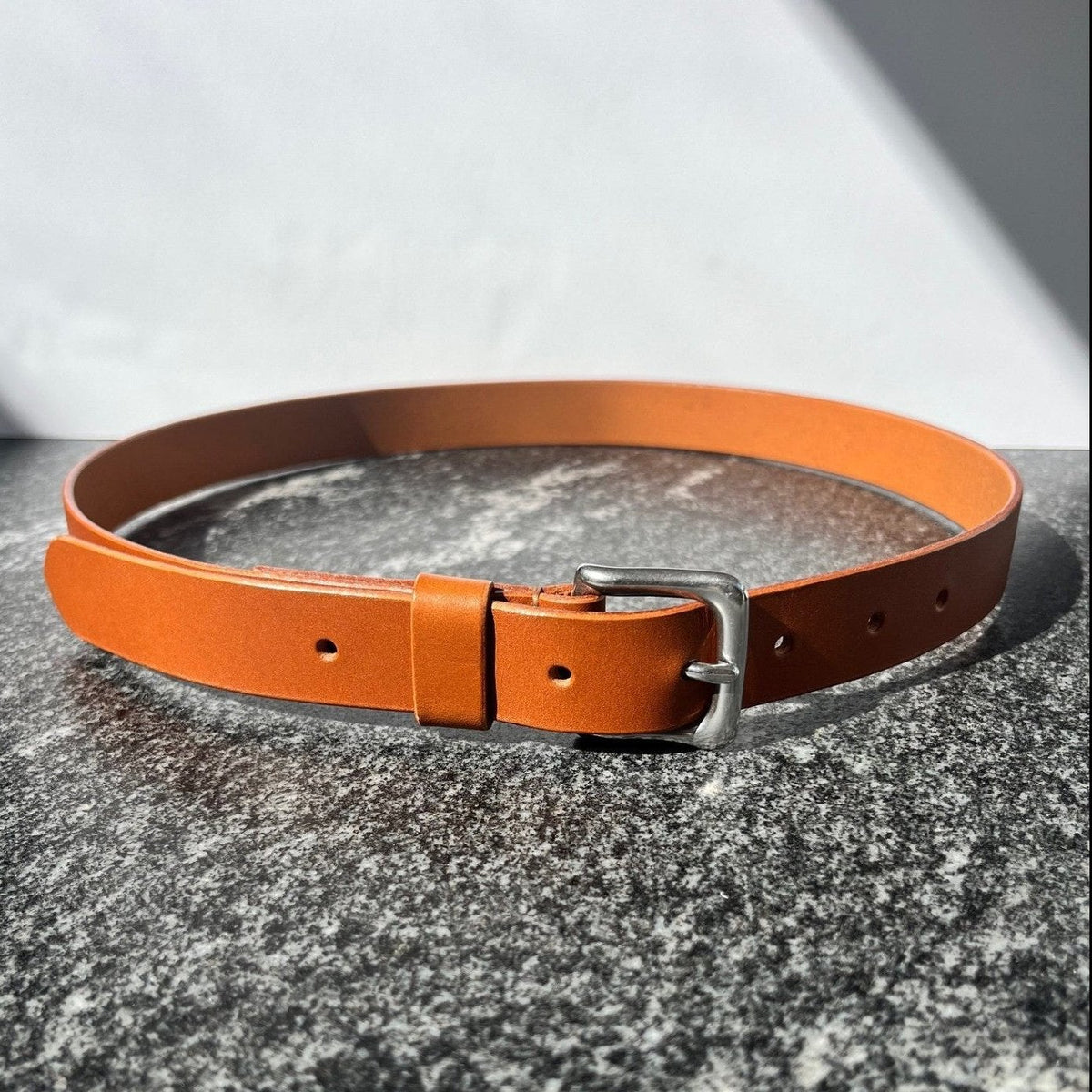 A tan leather belt with a silver-toned metal buckle, arranged in a circle on a textured gray stone surface with a white background.